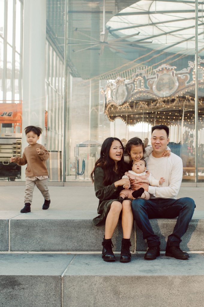 DUMBO Brooklyn NY is always so artistic, stylish and unique. Chic modern younger families, rich hipsters and even tourists love this place called DUMBO! Sally and Terence drove from Long Island stowing three children in the car. Of course I know and expect to get a text saying “sorry, but we are running late” at 3pm the time we are suppose to meet. I arrive early to park, so I wait in the car for about 30mins. In front of Ice Cream Factory 1 Water Street I see Sally. Been 8yrs since I photographed their wedding…how time flies, how amazing life is. Their three children are just adorable but we start with bit of cranky mood from woken up from their naps in the car. We walked around DUMBO and it’s sunset, magical hour as our backdrop we walk, we run and hop around the field. Truly running around in a big field is all that kids really need during the photoshoot. Sally and Terence Dumbo Fall Family Session photographed by Karis from Hey Karis. Photograph is like freezing time, motion in print, movie turned into a book.