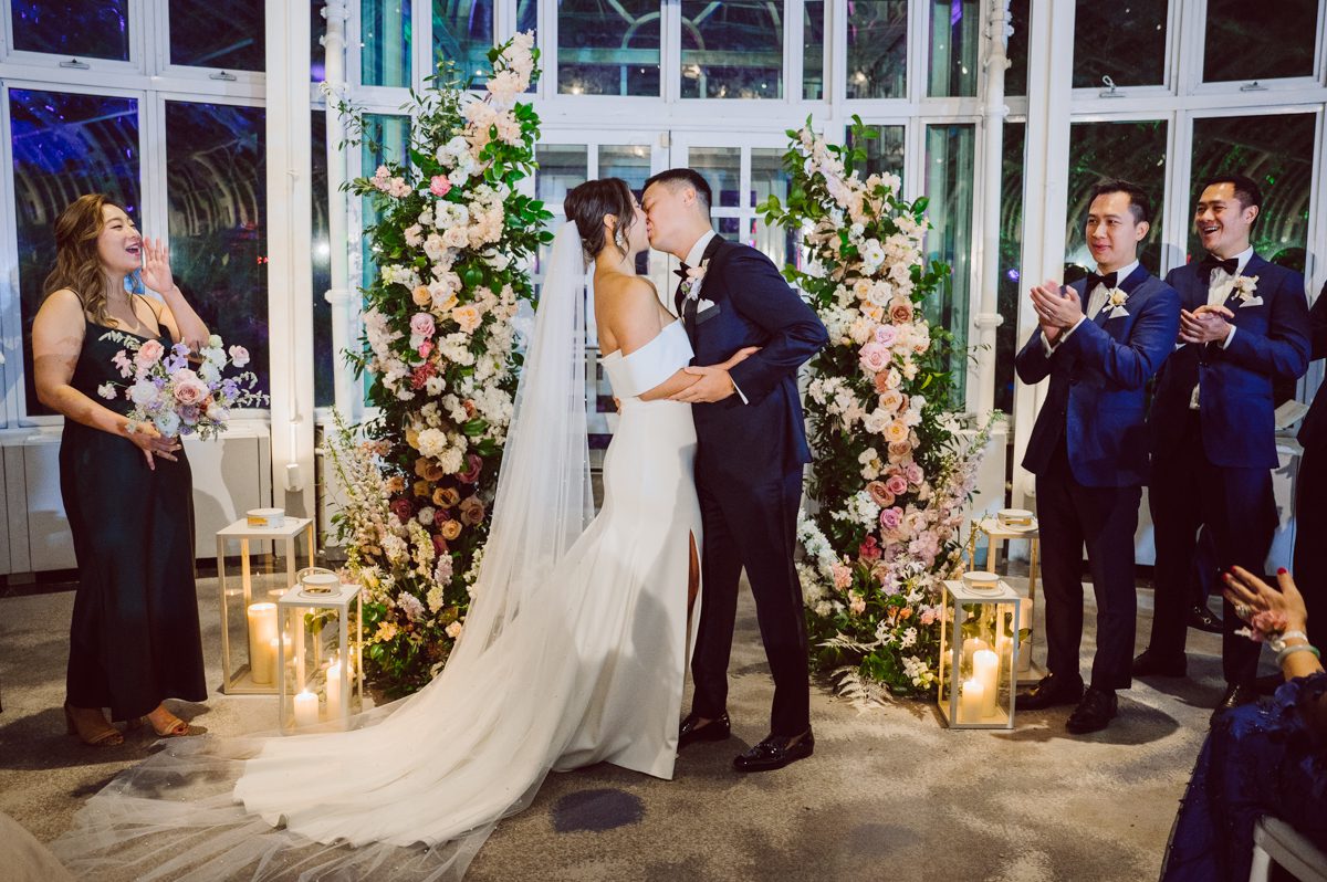 Duffield St, Brooklyn on November 19 even though it's Saturday morning it was still very busy. But luckily I found street parking right on Duffield St in front of Sheraton Downtown Brooklyn which was where I needed to be for Ariana and Jon's wedding prep. Vicky from VickyC5 hair/makeup team was there beautifying the ladies, I went straight to flat lays of Ariana's wedding details such as rings, jewelry and her wedding invitations. I use ribbons, extra loose flowers to make the flat lays more fine art looking. Once all the details are done, my full focus goes to the bride Ariana and her ladies. Ariana and Jon were planning to do a Chinese Door Game at the hotel room, so as soon as Ariana was ready in her Hanbok (Korean Traditional Wedding Outfit) Jon and his groomsmen arrived at the door. With Ariana's bridal suite crowded with too many people Ariana's bridesmaids started to give instructions to the games that Jon and his groomsmen need to play in order to get through the second door so that Jon can meet/see his bride Ariana. While the games are happening outside Ariana's door one of the bridesmaid has her phone on FaceTime so that Ariana can see how the men are doing during all the door games. I of course had my second shooter inside the room with Ariana to get her reactions which was hilarious. After the door games, we then moved to a conference room of at Sheraton Downtown Brooklyn for Ariana and Jon's Chinese Tea Pouring Ceremony (this is Jon's Chinese side's wedding tradition where you pour tea to all the elders in the family). Once all the Chines and Korean wedding traditions are over, we all rushed to Brooklyn Botanical Garden. Traffic from Sheraton Downtown Brooklyn to Brooklyn Botanical Garden was pretty bad, as usual. Once I arrived there of course was no street parking available near the entrance and parking lot for Brooklyn Botanical Garden was way too far for me to carry all the gears. After driving around I found one spot few blocks away from the Brooklyn Botanical Garden entrance. Brooklyn Botanical Garden was busy with Lightscape preparation and installation which we were not allowed to photograph so we were left with very little options for wedding portrait backdrop, plus it was COLD and we were looking light! These are important things to consider when you are planning for Winter wedding. I worked as quickly as possible with Ariana and Jon's first look and bridal party photos. Then we stayed inside for family photos since elders did not wanted to go outside which I totally don't blame. Then I quickly took Ariana and Jon outside for bit more couple's portraits then it was time for the wedding ceremony. Ariana and Jon's wedding ceremony held place at The Palm House at Brooklyn Botanical Garden, but because it was an winter wedding we've lost all the light and I had to use my video lights but over all the ceremony was beautiful and Oh Fleur NYC did absolutely an amazing job with ceremony decor (floral/candles). The Palm House was empty again, OhfleurNYC as well as Brooklyn Botanical Garden staff were busy turning the room. Oh Fleur NYC's Jessica's talent was amazing her usage of floral, choice of colors and mixing the floral with tall candles were magical. Turned The Palm House into a beautiful and romantic garden. Warmth of candles, peach and pinks of flowers all blended so well turning the Palm House at Brooklyn Botanical Garden in to fairytale floral ballroom. Ariana and Jon attended cocktail enjoying their time with friends and family before their wedding reception at The Palm House. Ariana and Jon's wedding at The Palm House at Brooklyn Botanical Garden documented by Karis from Hey Karis New York, Brooklyn, Hudson Valley and Catskills Wedding Photographer and NYC Elopement Photographer.