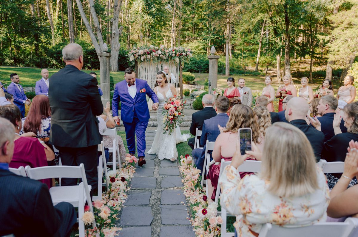 How do I explain the sincere and true feelings that Izzy and Brendan shared on their wedding day at one of the most magical place Arrow Park in Monroe NY. Izzy is Opera singer and Brendan is a Chef who owns his own restaurant called Circle in Newton, NJ got married at Arrow Park NY a month ago and it got real festive and wild real fast. But let's come back to the wild party in a bit and talk about their morning first. Izzy and Brendan both got ready onsite at Arrow Park, great thing about Arrow Park is that it has enough rooms to host all their bridal party and family. With all of Izzy and Brendan's loved ones onsite Izzy and Brendan's wedding prep was fun. No drama and no one running late, Izzy and Brendan's wedding prep went so smooth with everyone's cooperation! Izzy got into her dress with her mom and sister by her side, then we went down to the back steps for Izzy and Brendan's first look. Oh boy Brendan got super emotional for their first look, he couldn't even turn around to look at Izzy with all the happy emotions rushing to him. Izzy held his hand and they looked at each other. The two of them cried. Sharing such a beautiful first look Izzy and Brendan walked out to the front of Arrow Park for group and family photos. It was an August wedding so I kept everyone in the shade not too far, making it simple easy for group and family photos are important job to wedding photographer. After all the formal group photos were done, it was almost time for their wedding ceremony at the garden. Beautiful decor and floral arrangements were set up in the garden ready for Izzy and Brendan's wedding ceremony, guest started to walked down the long driveway and down the steps and found seats waited for the bride and groom to walk down the aisle. Brendan now standing in the front next to the priest, started tearing up as soon as he saw Izzy coming down the aisle escorted by her father. The two linked their eyes at each other and in the most loving way possible looked at each other through out the ceremony then sealed it with the first kiss as husband and wife. As calm and peaceful as Izzy and Brendan's wedding ceremony was, the two entered their reception each with (shaken) champagne bottles in their hands, everyone and anyone in Izzy and Brendan's way got sprayed with Champagne! They jumped, pouring Champagne into their friends mouth straight out of the bottle, then each drank some out of the bottle. Building the excitement in to their epic wedding party, Izzy and Brendan held each others hands and walked into the center of Arrow Park's courtyard and danced their first dance. Smoke and smell of freshly cooked dinner being prepared by Brendan's friends at Promises Kept Hudson Valley NY was filling the air, as first courses were being served. Happiest sound of plates, utensils and people chatting, Izzy and Brendan sat at their sweetheart table to enjoy their wedding dinner prepared by Promises Kept Hudson Valley NY. Then the evening got deeper and darker and vibe got stronger and crazier. Music got louder and everyone was in their element celebrating Izzy and Brendan on their wedding day. Izzy and Brendan's Arrow Park Monroe NY Wedding documented by by Karis | Hey Karis | Catskills, Hudson Valley NY elopement and wedding photographer | Brooklyn NY elopement and wedding photographer | Manhattan NY elopement and wedding photographer | Hey Karis is fun documentary wedding and elopement photographer.